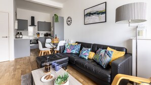 Een moderne woonkamer met een zwarte leren bank, een salontafel met gebak, een eethoek met stoelen en een keuken op de achtergrond.