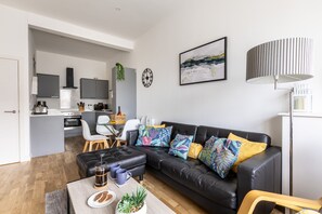 A modern living space with a black leather sofa, a coffee table, and a kitchen area in the background.