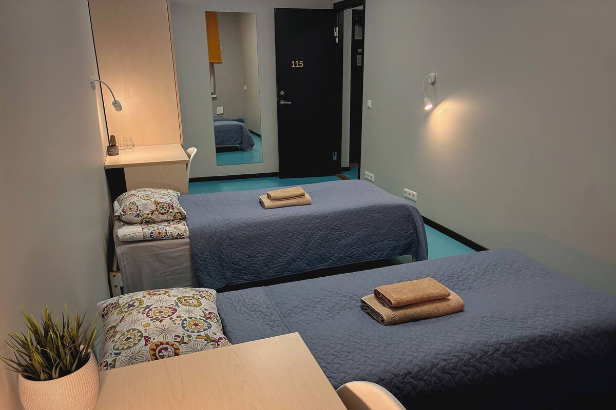 Habitación de hotel con dos camas, una mesita, una planta en maceta y una puerta con el número 115.
