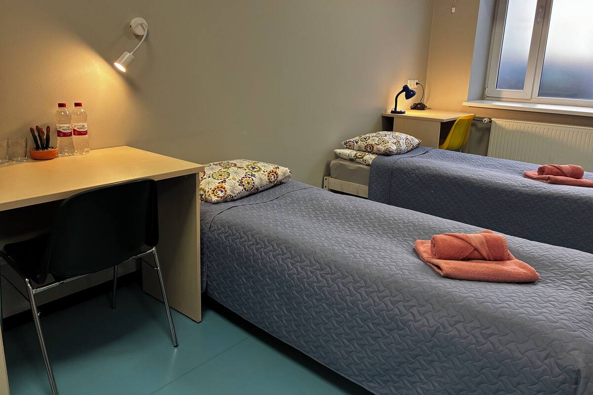 Una habitación con dos camas, un escritorio, una silla, una ventana y una mesita de noche con lámpara.