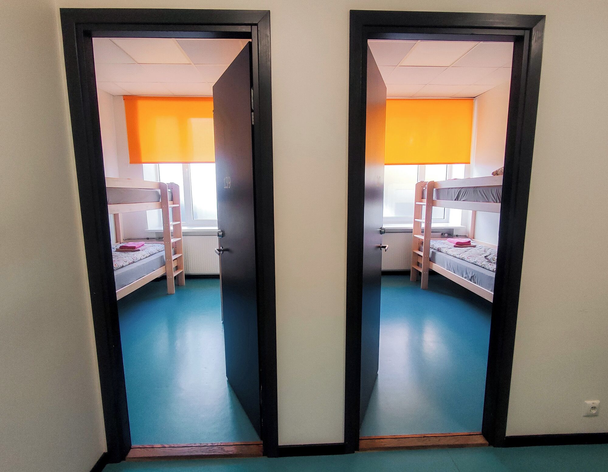 Habitación con espejos, literas, una ventana con persiana naranja y una puerta con el número 139.