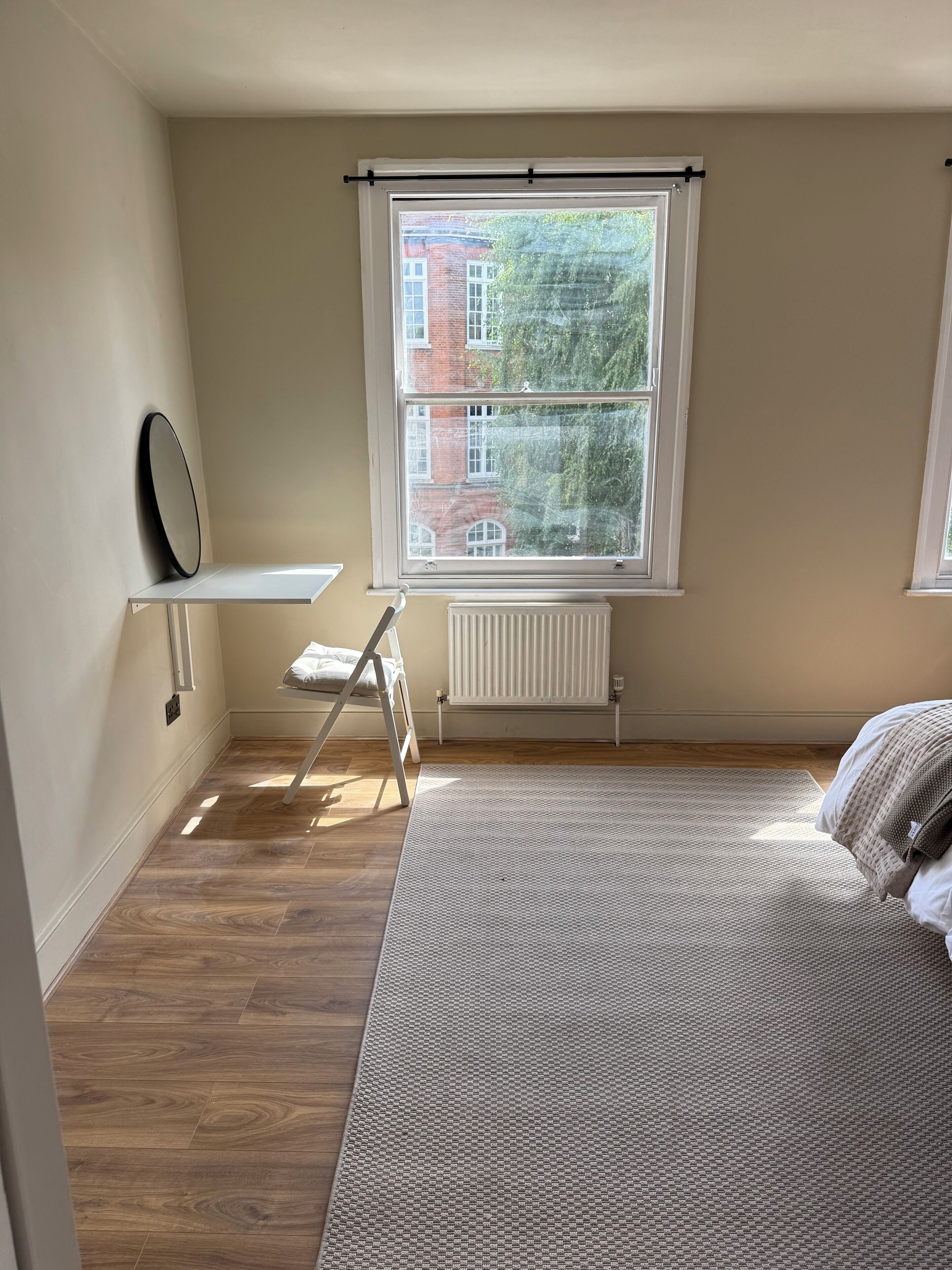 A bedroom with a bed, a window, a radiator, and a shelf with a mirror.