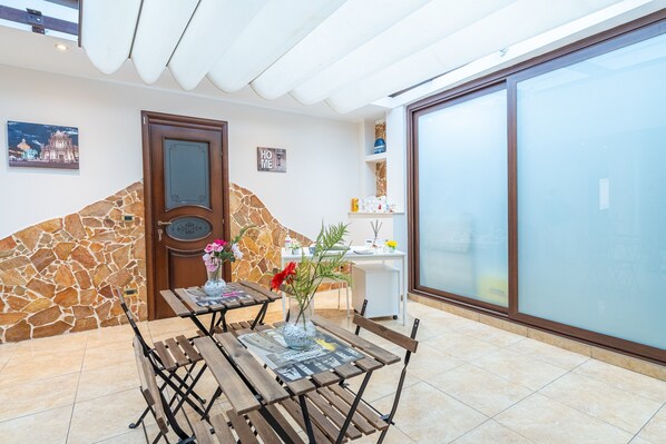 A room with a stone wall, a wooden door, a table with chairs, a vase with flowers, and a large glass door.