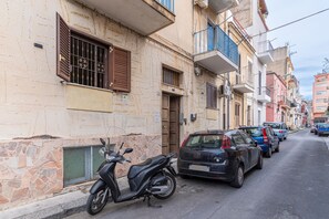 A narrow street with parked cars and a scooter, buildings with balconies, and a stone facade.