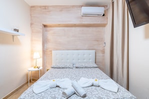 A hotel room with a bed, a bedside table with a lamp, a TV mounted on the wall, and an air conditioning unit.
