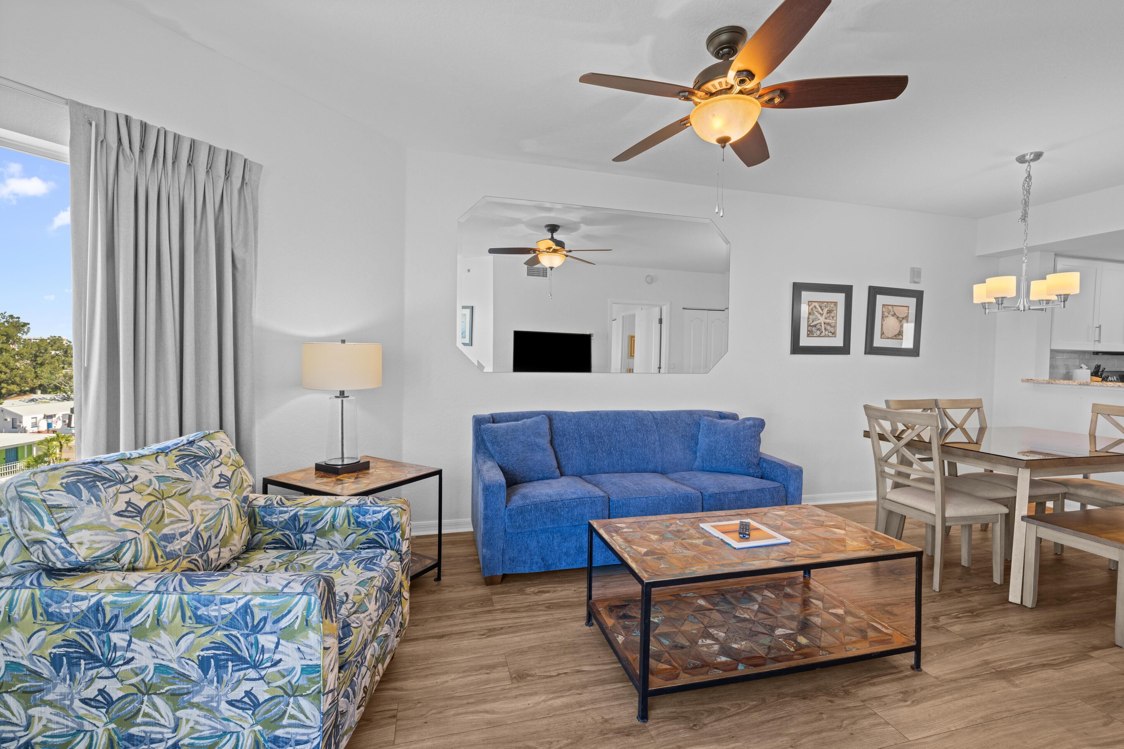 Ein modernes Wohnzimmer mit einem blauen Sofa, einem gemusterten Sessel, einem Couchtisch, einem Deckenventilator, einem Fernseher und einem Essbereich mit Stühlen und einem Tisch.