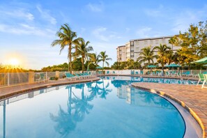 Una piscina con sillones de descanso y palmeras, rodeada por un edificio.