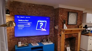 Una habitación con pared de ladrillo, un televisor de pantalla plana mostrando un juego de preguntas y respuestas, un mueble de entretenimiento azul y una chimenea.