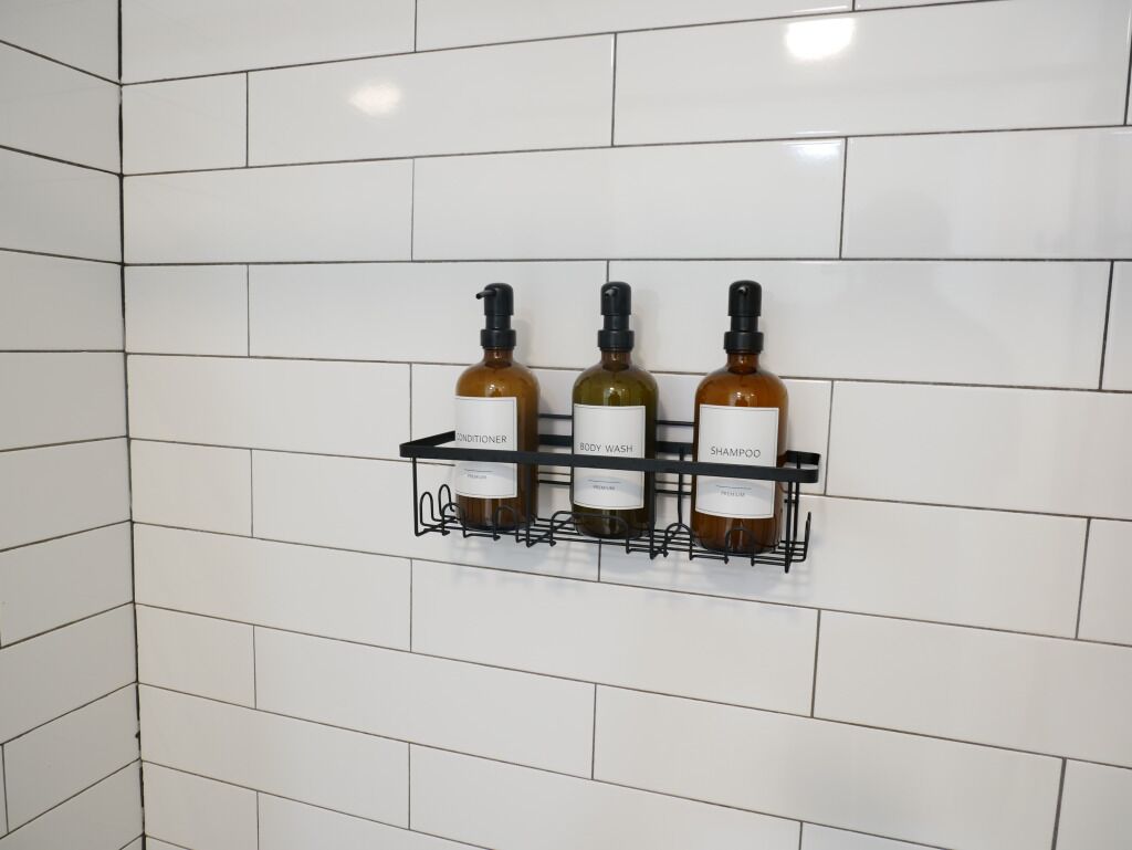 Un baño con paredes de azulejos blancos y un estante negro que contiene tres botellas etiquetadas como acondicionador, jabón corporal y champú.