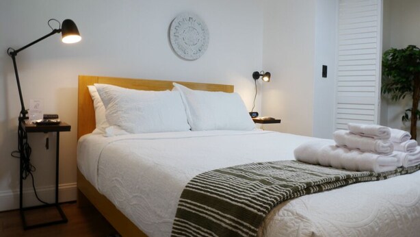 Una cama bien hecha con sábanas blancas, una lámpara de noche y una toalla doblada al lado.