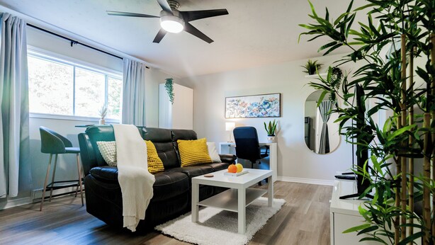 Una sala de estar con un sofá de cuero negro, una mesa de centro blanca, un sillón verde y un ventanal con cortinas.
