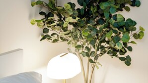 Una cama bien hecha con ropa de cama blanca, una mesita de noche blanca con una lámpara dorada y una planta artificial montada en la pared.