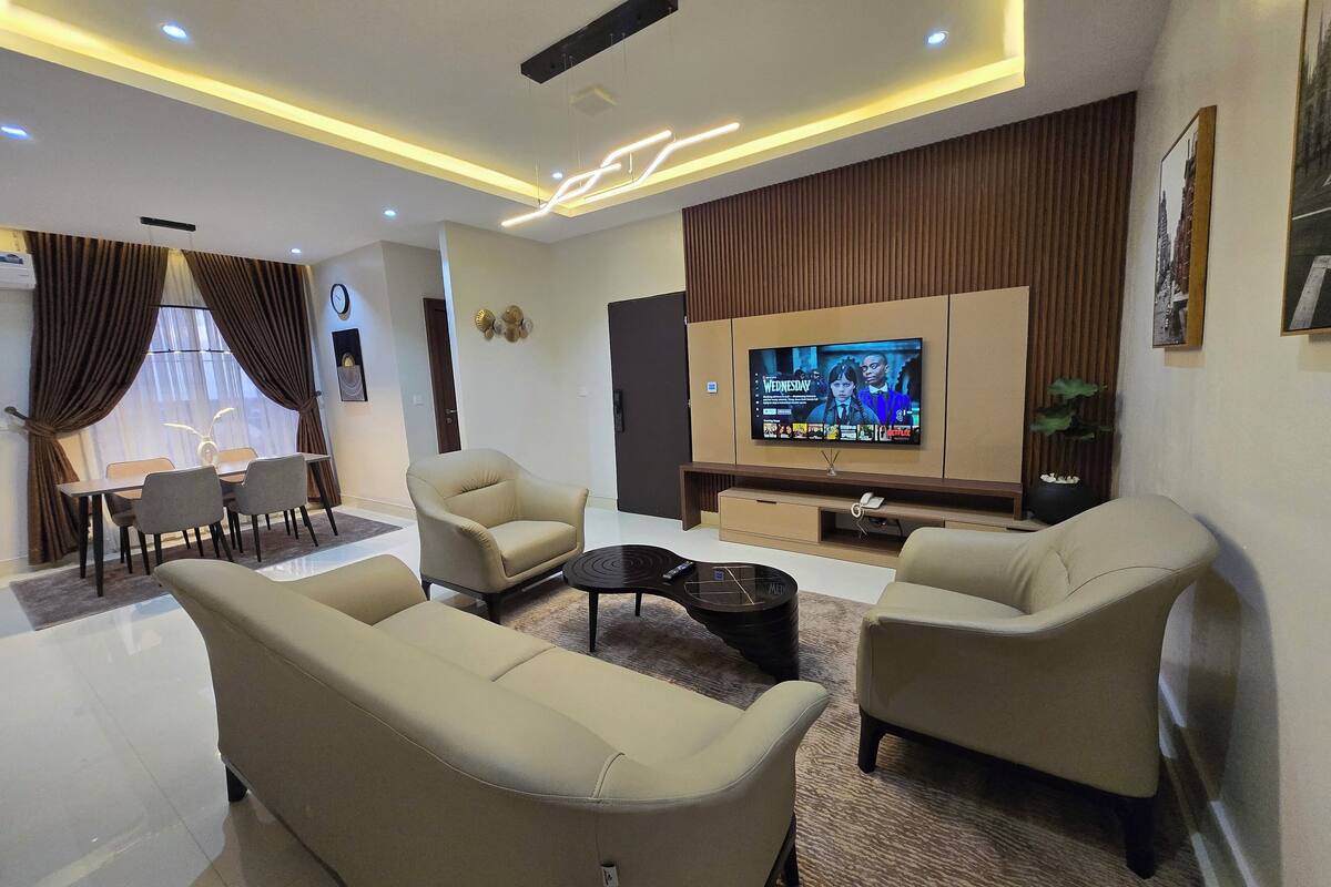 A modern living room with a flat-screen TV, beige sofas, a coffee table, and a dining area with a table and chairs.