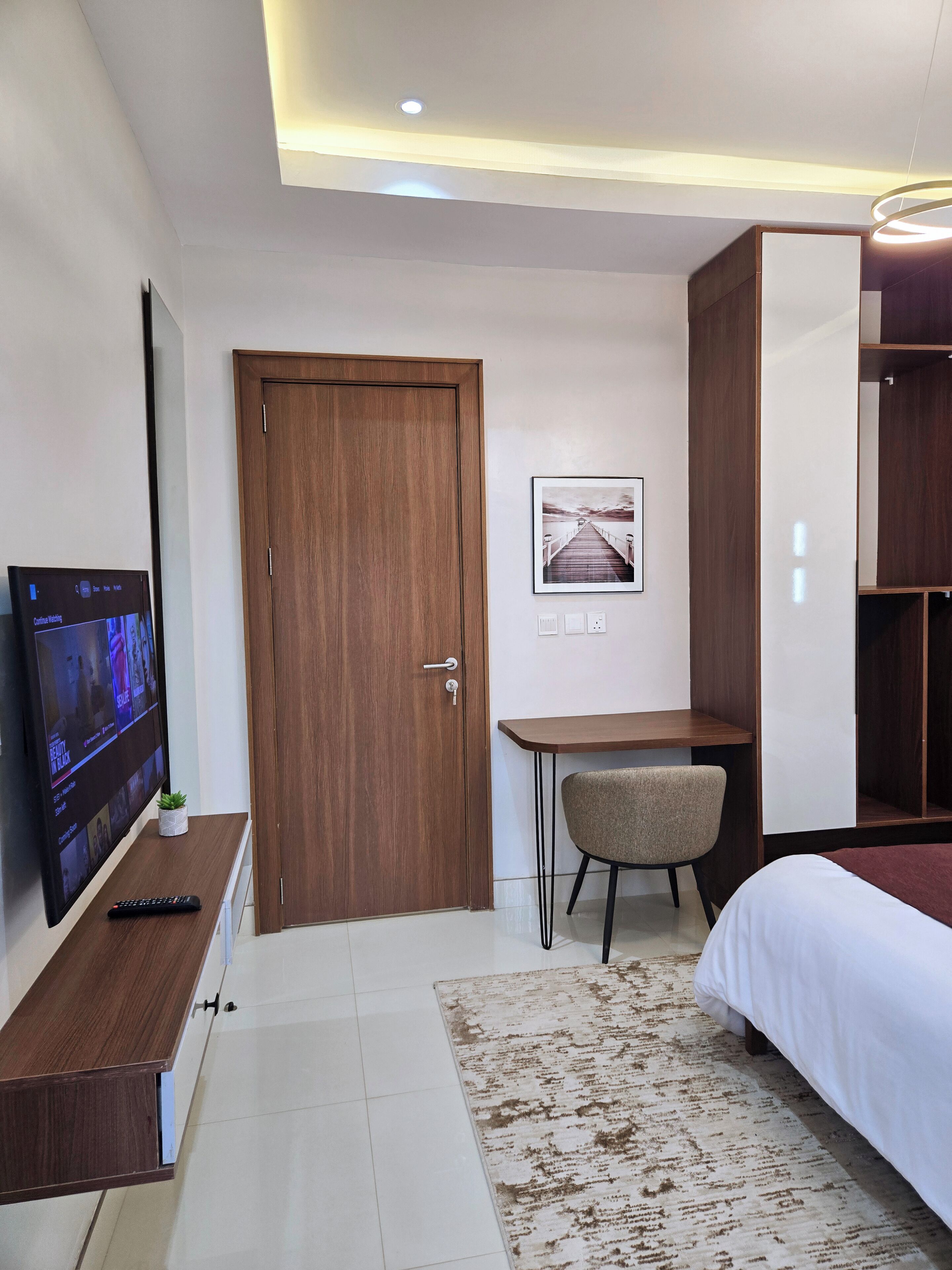 Habitación de hotel con puerta de madera, un escritorio con silla, cama, televisor sobre un mueble de madera y un cuadro en la pared.