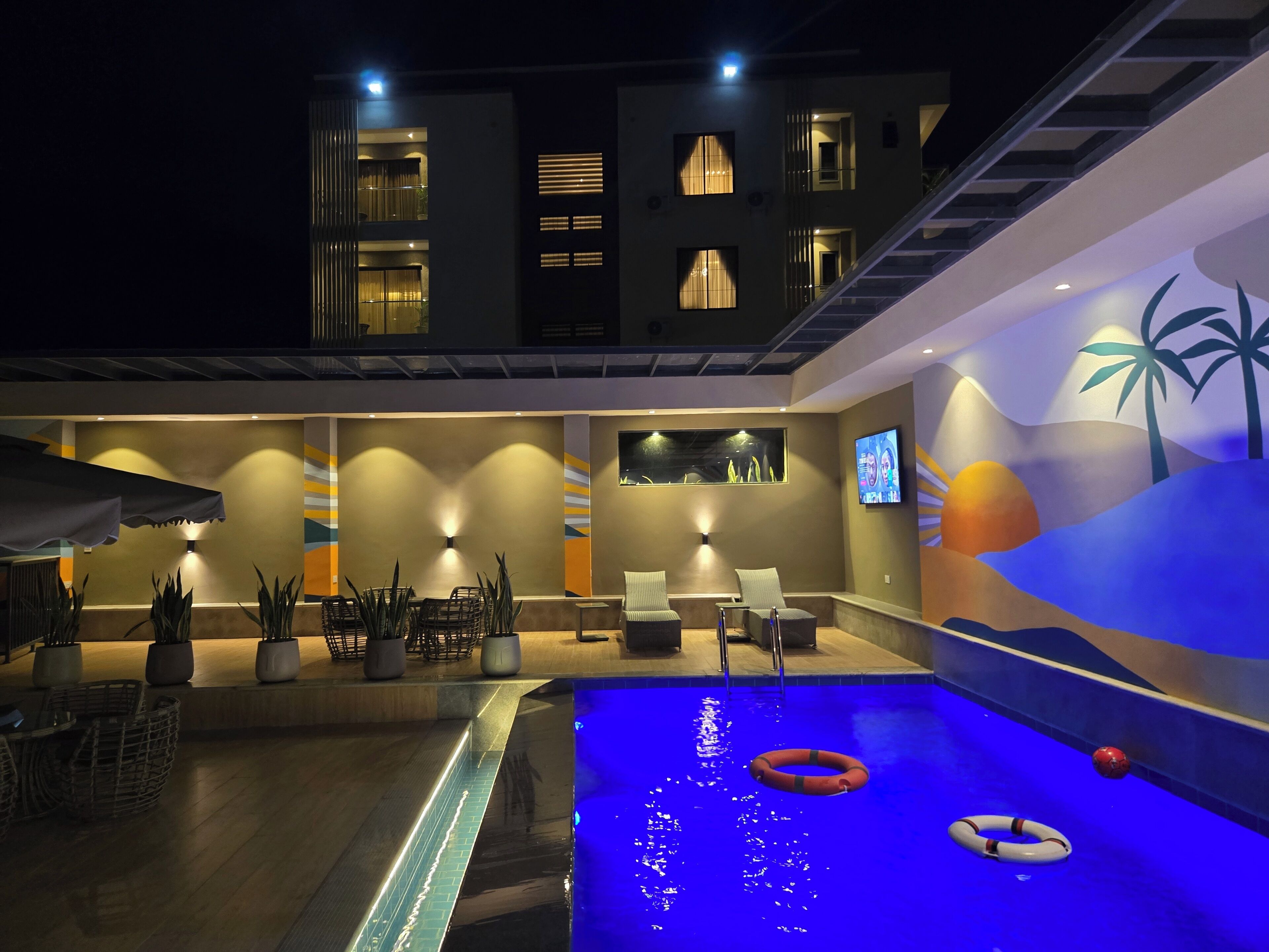 A swimming pool with two lifebuoys, a lounge area with chairs and tables, and a TV screen on the wall.