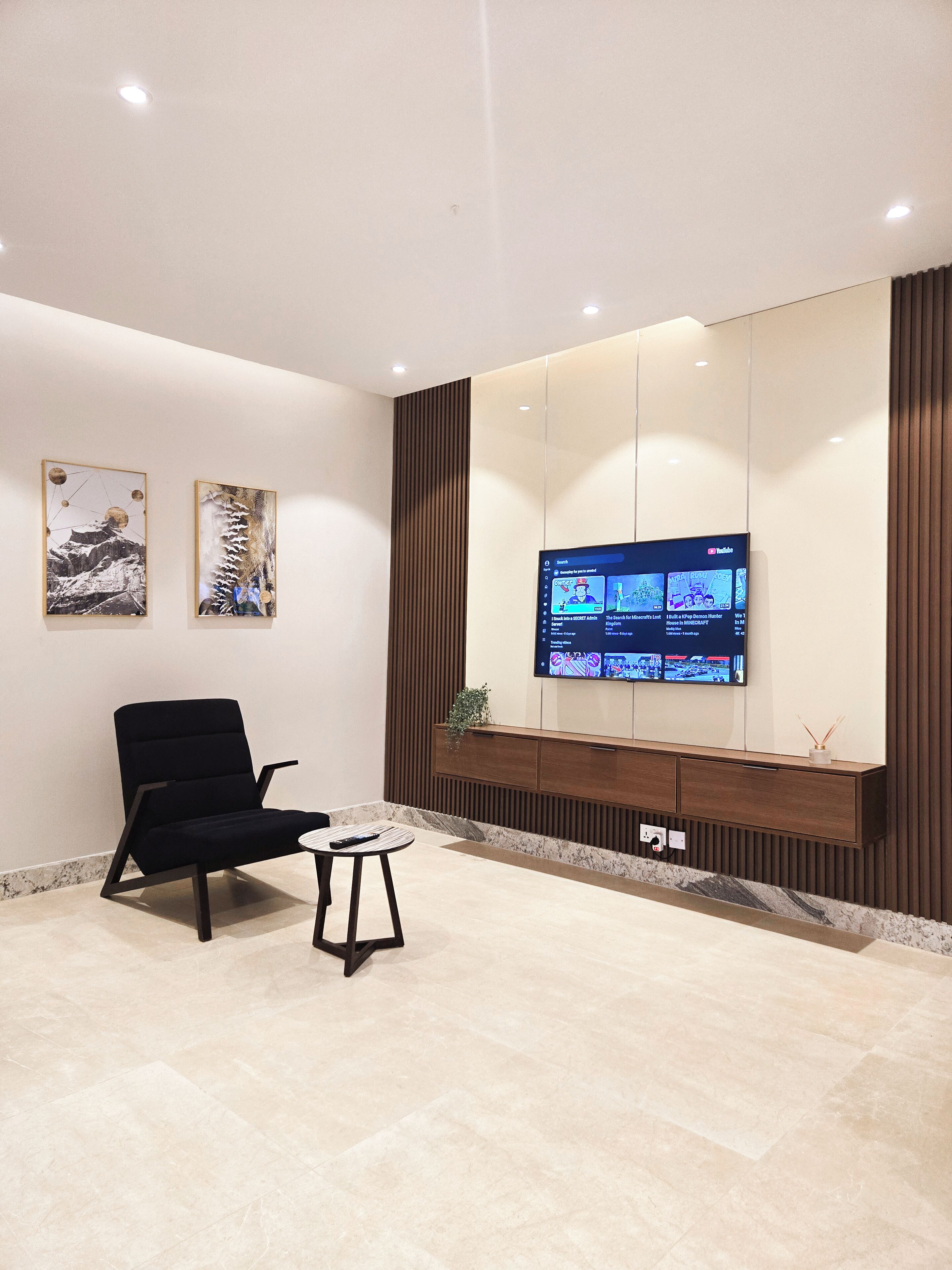 A modern waiting area with a black chair, a small table, and a flat-screen TV mounted on the wall displaying various channels.