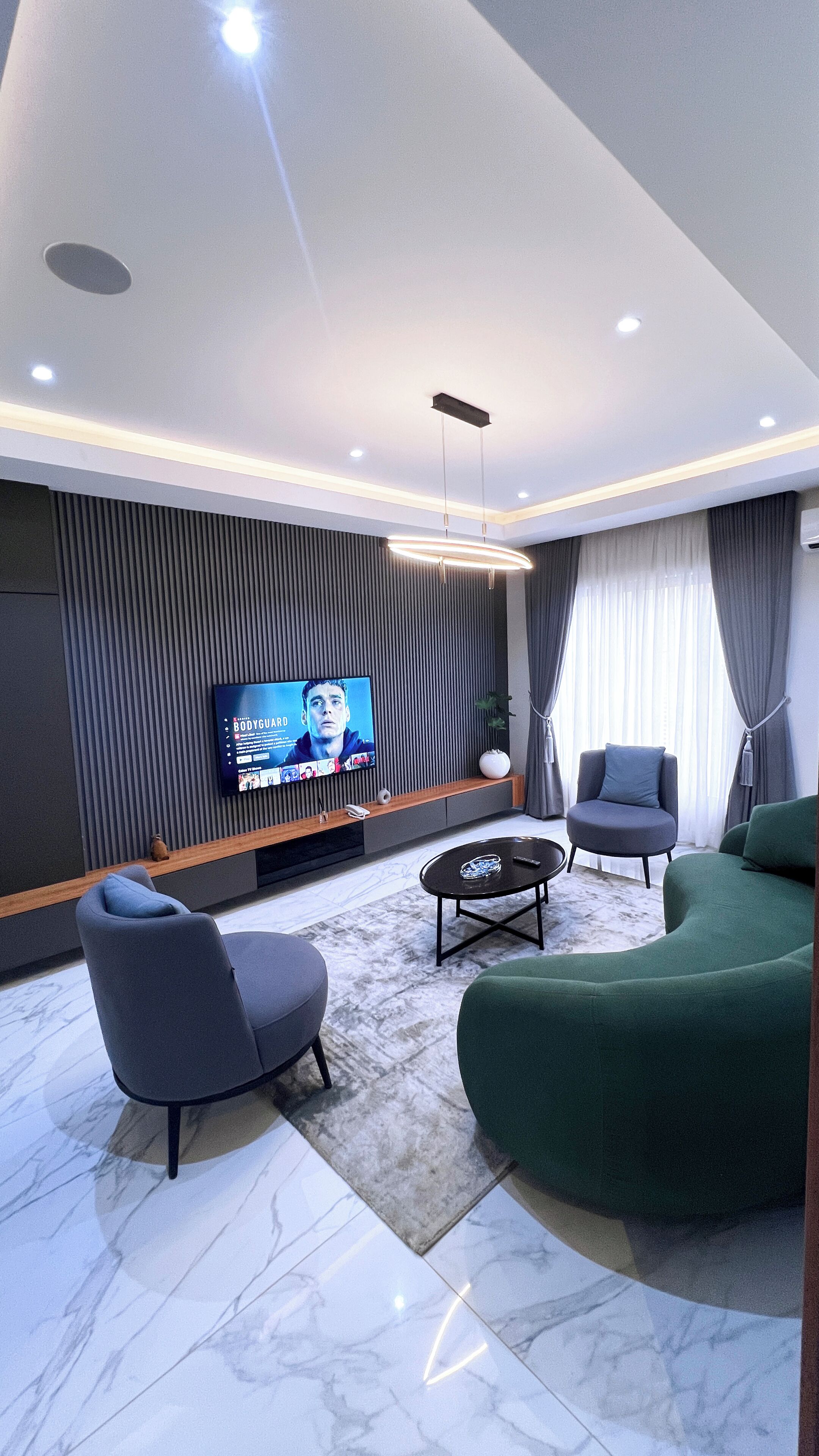 Ein modernes Wohnzimmer mit Flachbildfernseher, zwei Sesseln, einem grünen Sofa und einem Marmorboden.