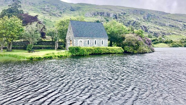 A stone building with a sloped roof, situated by a body of water, surrounded by lush greenery and hills.