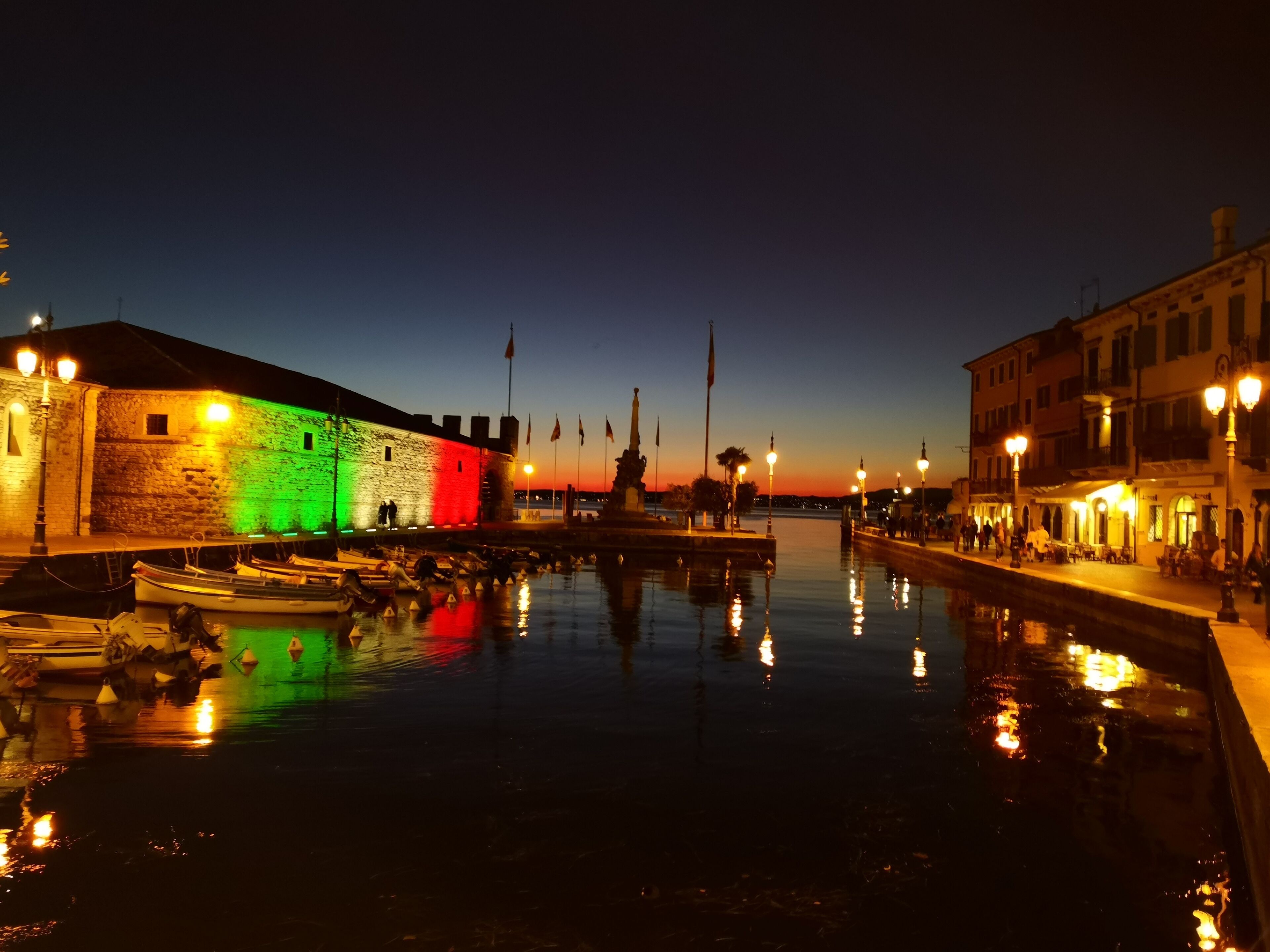 Een watergezicht met een stenen gebouw dat verlicht is door kleurrijke lichten, een kalm waterlichaam dat de lichten weerspiegelt, en een heldere lucht.