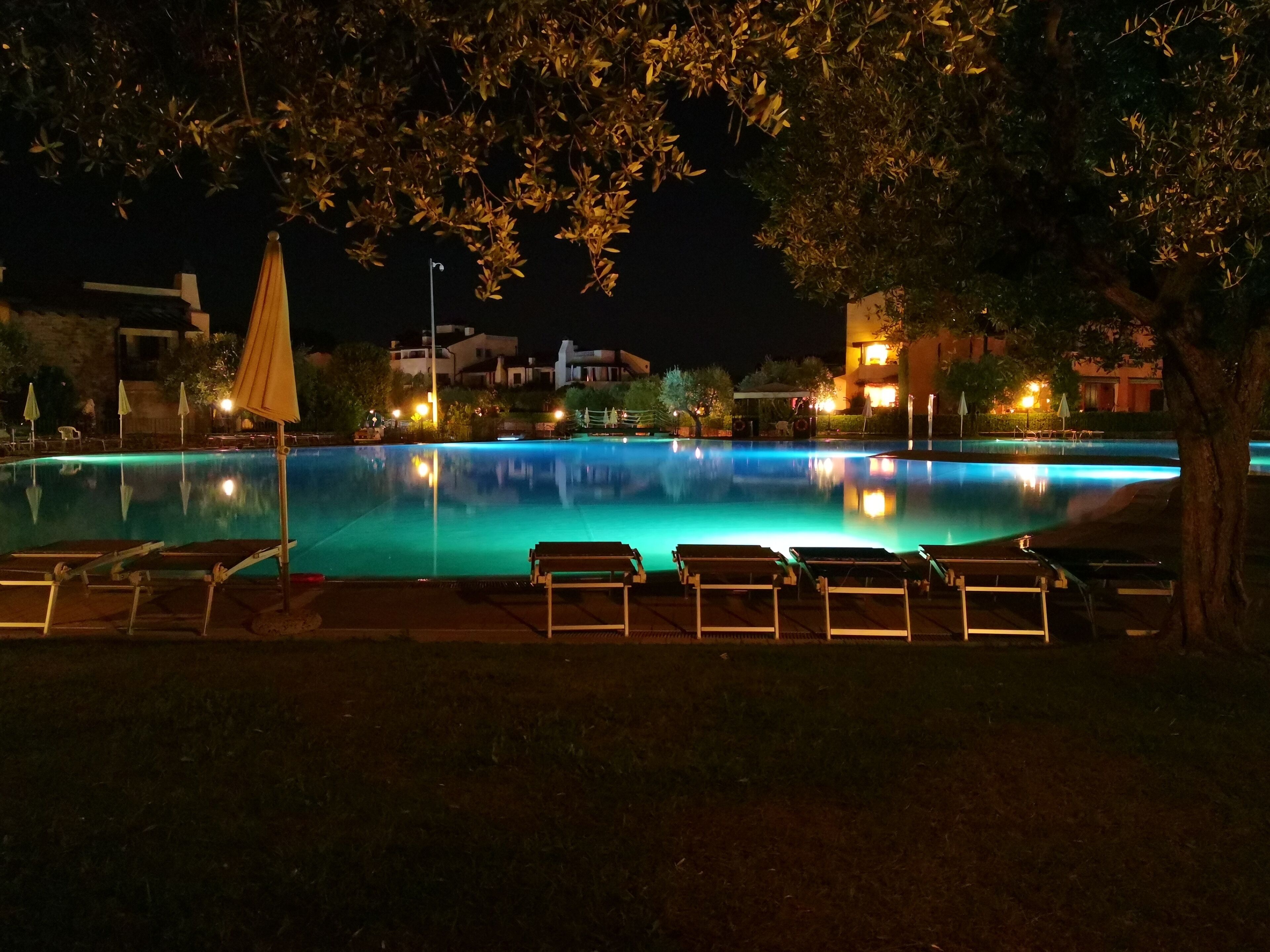 A nighttime pool area with lounge chairs and umbrellas, surrounded by buildings and trees.