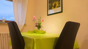 A dining area with a table covered in a green tablecloth, two black chairs, a vase with flowers, and a framed picture on the wall.