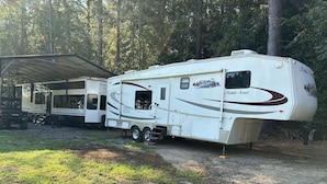 A mobile home with a covered porch and a trailer attached, parked in a wooded area. - Welcoming RV with AC in charming Farmerville. (Farmerville)