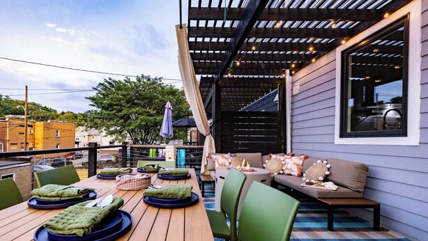 An outdoor patio with a wooden table set for dining, green chairs, and a cushioned seating area with a fire feature.
