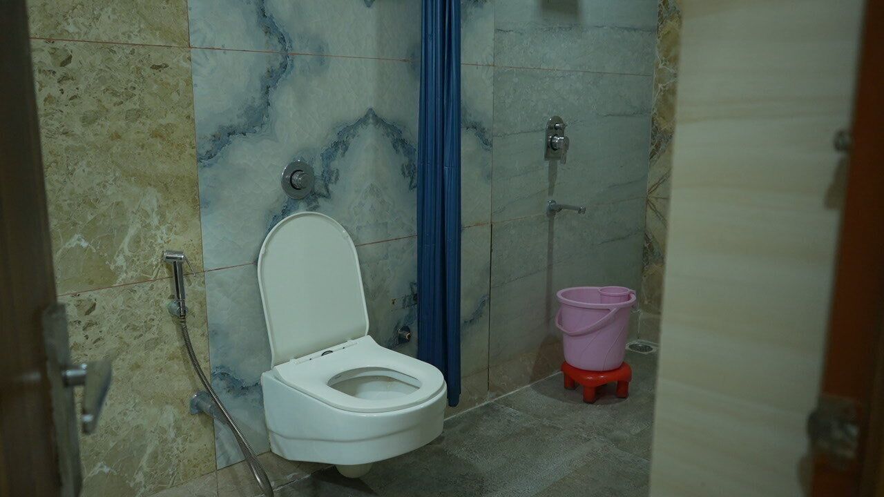Une salle de bain avec des toilettes blanches, un rideau de douche bleu et un seau rose posé sur un tabouret rouge.