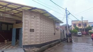 Un edificio con entrada de baldosas, una zona cubierta con techo metálico y un muro de ladrillo.