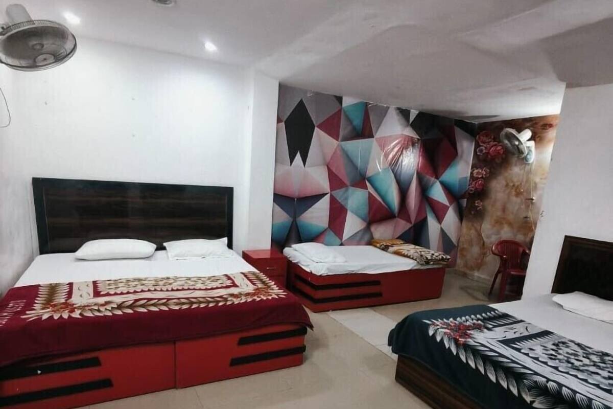 A hotel room with two beds, a red and a blue bedspread, a geometric wall mural, and a ceiling fan.