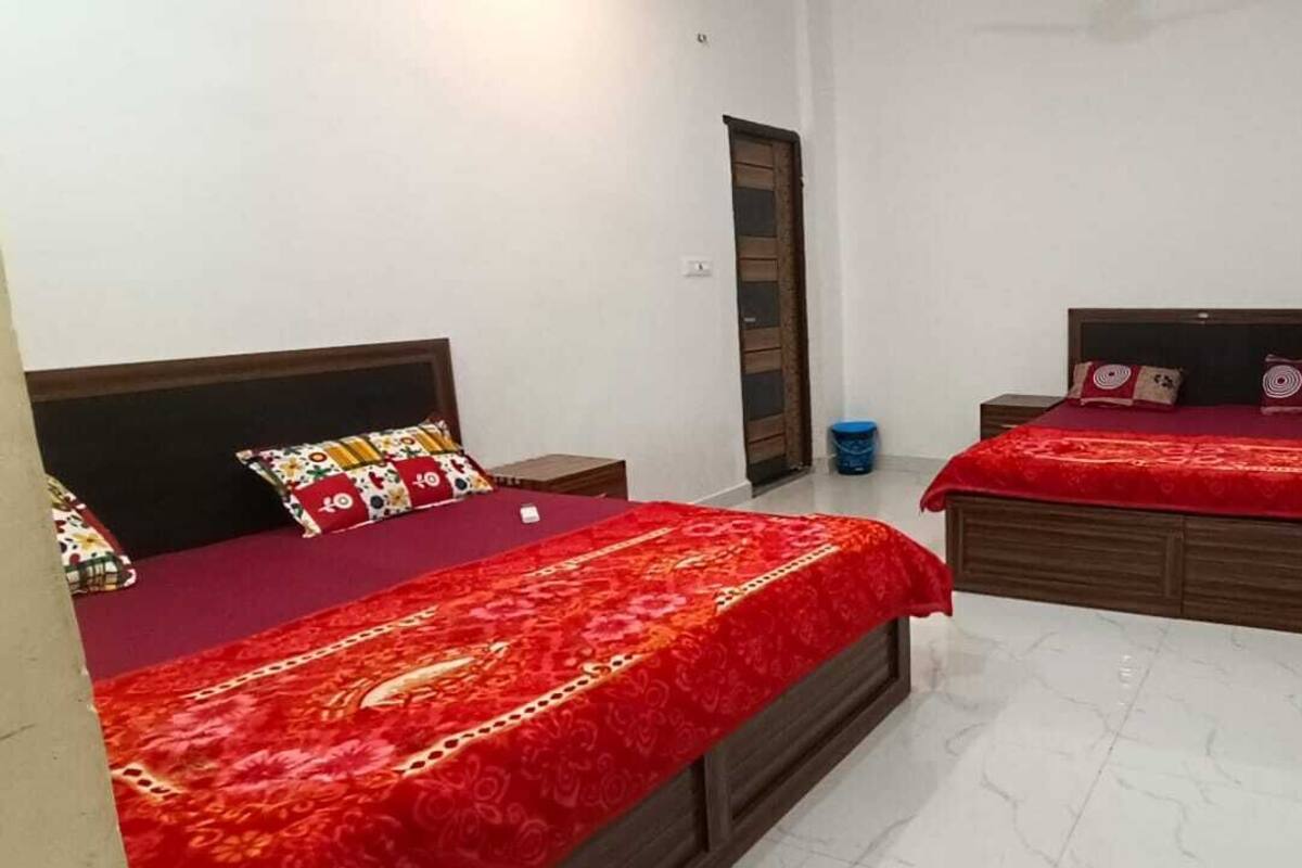 A small, clean room with two beds, each with a red bedspread and matching pillows. There is a wooden door on the right and a blue trash bin against the wall.