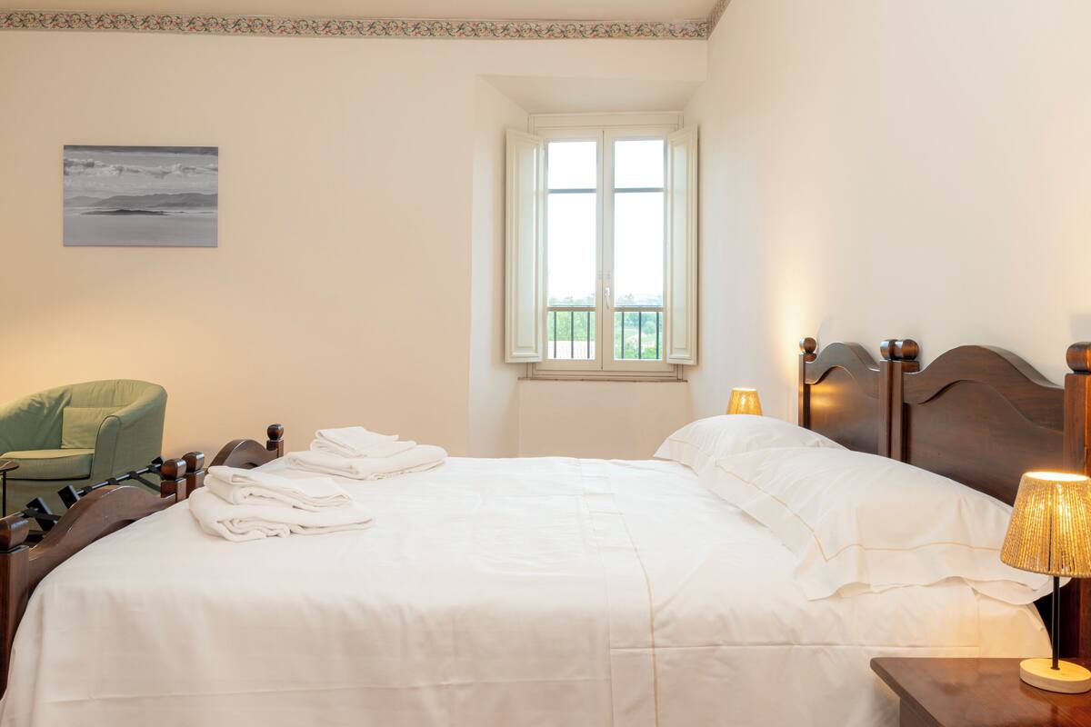 A neatly made bed with white linens, a green armchair, a window with a view, and a lamp on the bedside table.