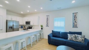A modern kitchen with white cabinets, a stainless steel refrigerator, and a microwave. There are white stools and a blue sofa with patterned cushions. - Coastal Beach House, Blocks from Ocean! (Myrtle Beach)