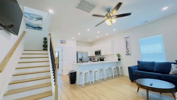 A modern kitchen with white cabinets, a central island with bar stools, a ceiling fan, and a large window with blinds. - Coastal Beach House, Blocks from Ocean! (Myrtle Beach)