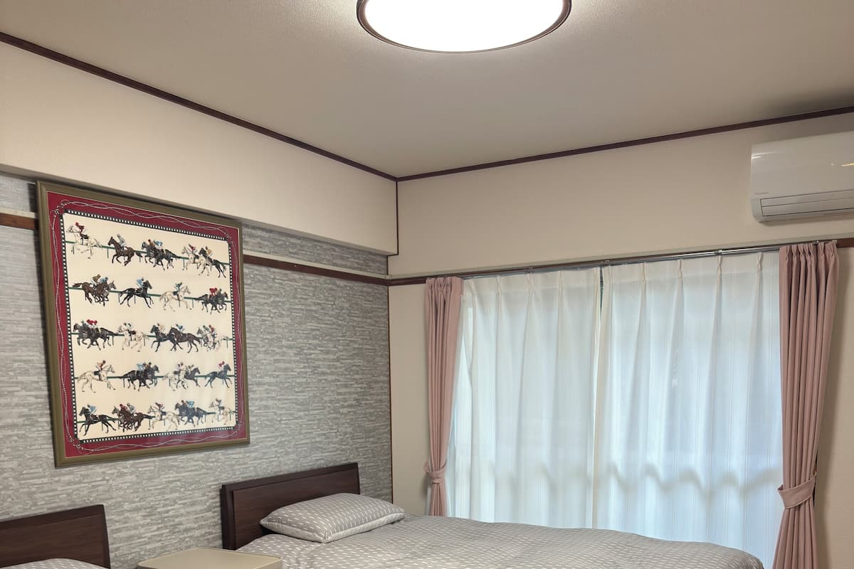 Une chambre d’hôtel avec deux lits, une petite table, un climatiseur fixé au mur et une grande fenêtre avec des rideaux.