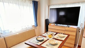A modern living room with a sofa, a dining table set for two, a flat-screen TV, and a ceiling-mounted light fixture. - HYGGE HOUSE Traveler's house (Kashiwa)