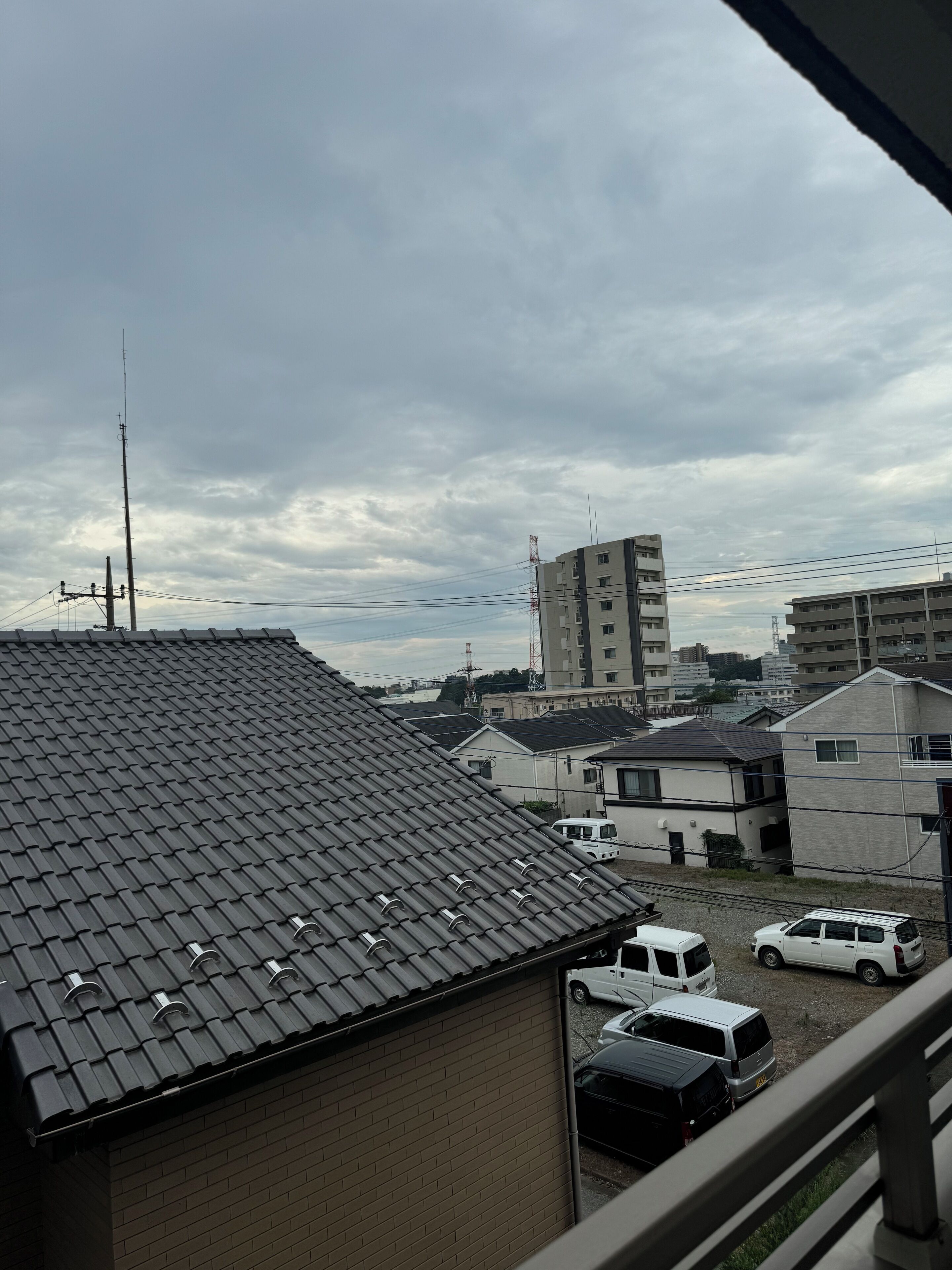 Una zona residencial con varias casas, algunas con tejados de tejas y otras con tejados planos. Se ven varios coches estacionados y una torre de líneas eléctricas al fondo.