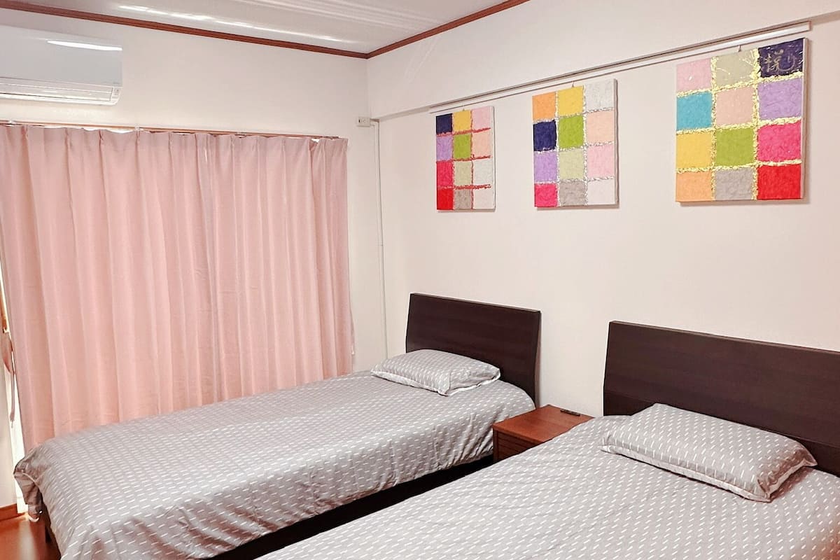 Une chambre d’hôtel avec deux lits simples, un luminaire au plafond, un appareil de climatisation et une œuvre d’art colorée accrochée au mur.