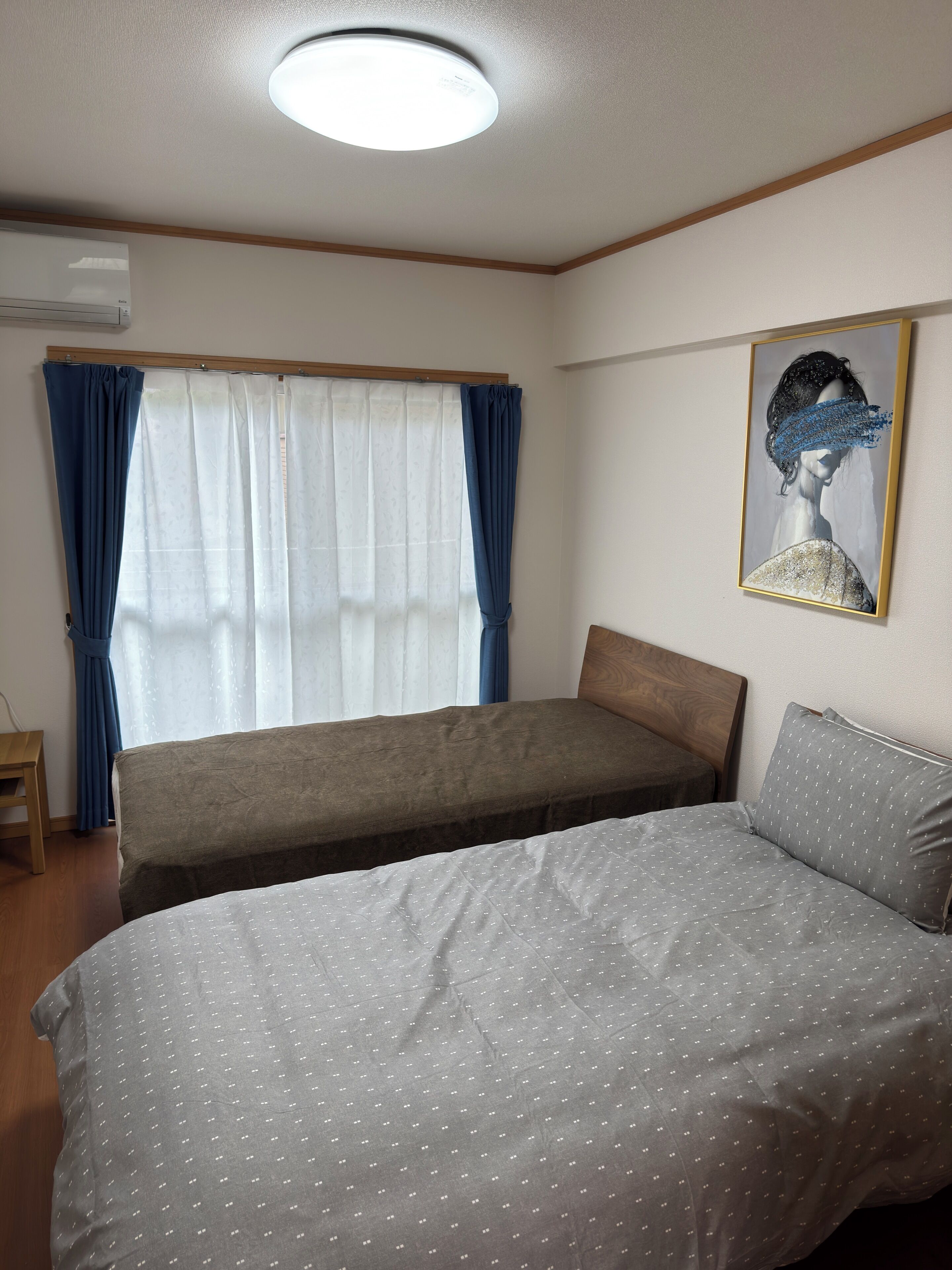 A room with two beds, a window with curtains, a ceiling light, and a framed artwork on the wall.