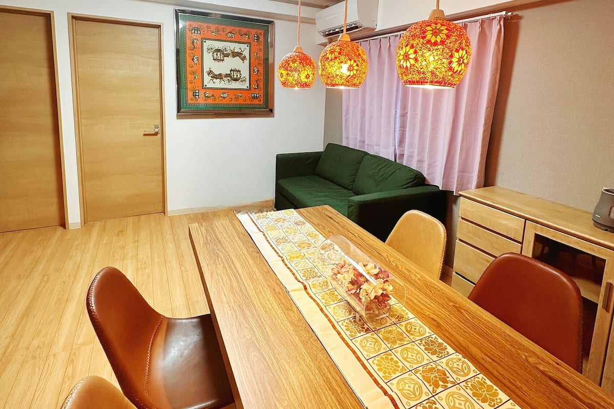Un espace repas comprenant une table en bois, des chaises en cuir marron, un canapé vert et des lampes décoratives suspendues.