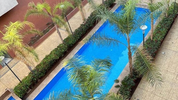 A rectangular swimming pool surrounded by palm trees and greenery, with a tiled walkway and a building in the background. - Sérénité Orientale I Apartment in Marrakech (Marrakesh)