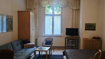 Ein möbliertes Zimmer mit einer Couch, einem Sessel, einem kleinen Tisch, einem Fernseher und einem Schrank.