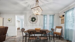 A dining area with a wooden table, six chairs, a chandelier, a wreath on the wall, and light blue curtains.