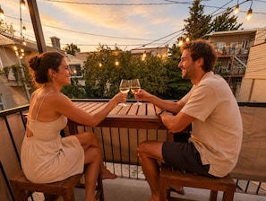 A couple seated at an outdoor wooden table with string lights overhead, surrounded by greenery and residential buildings.