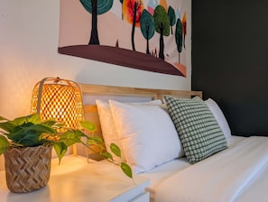 Un lit bien fait, avec des oreillers blancs et à motifs, un pot de fleurs en osier et une lampe fixée au mur.