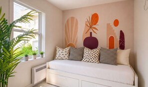 A bedroom with a bed, a radiator, a window with plants, and a wall with abstract art.