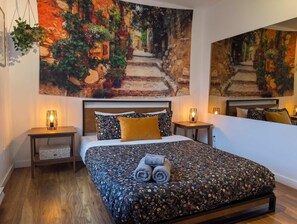 Une chambre à coucher comprenant un lit, deux tables de chevet, une commode, une grande couvre-lit à motifs floraux, une tête de lit et un grand tableau représentant une scène de jardin accroché au mur.