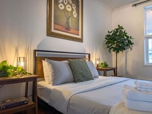 Una cama bien hecha con sábanas blancas, un almohadón verde y una mesita de noche con una lámpara y una planta.