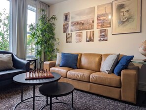 Una sala de estar con un sofá de cuero marrón, una mesa redonda de centro y un tablero de ajedrez. Hay cuadros en la pared y una planta en la esquina.