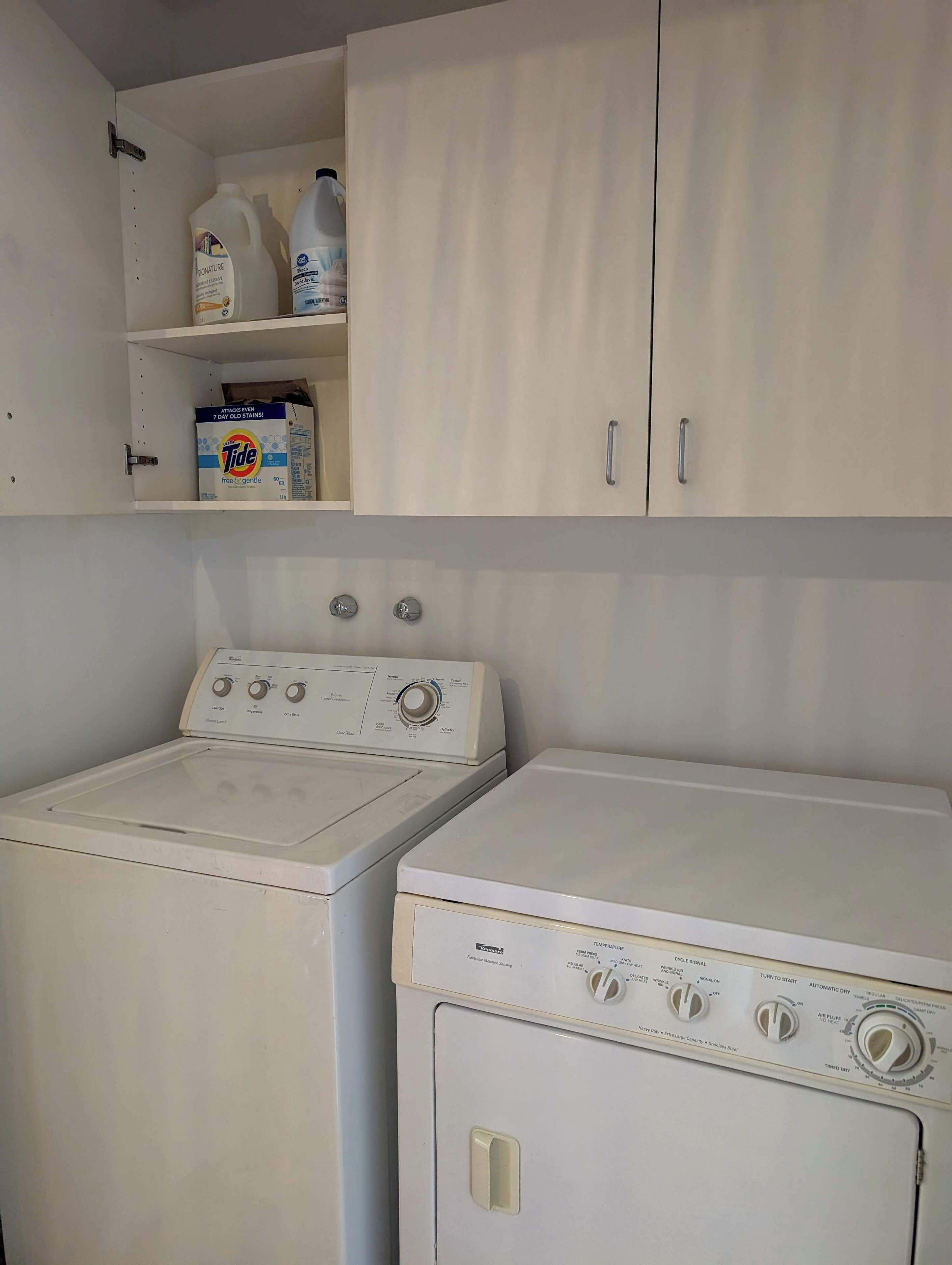 A laundry area with two washing machines, a shelf with detergent bottles, and a cabinet above.